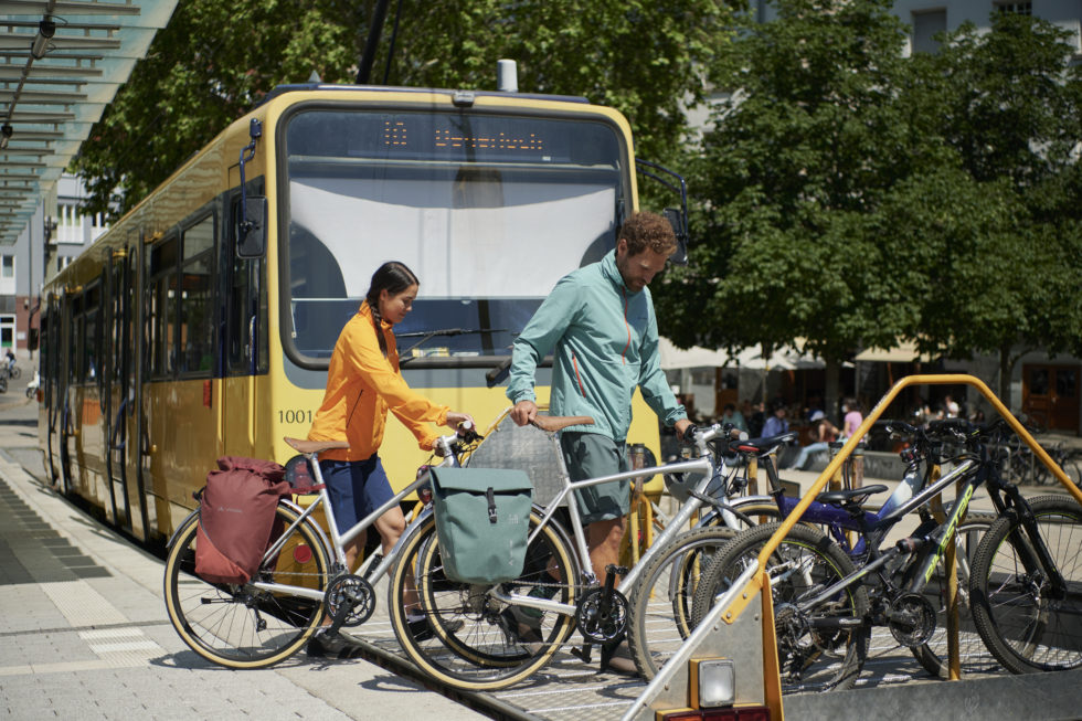 Ein Mann und eine Frau stellen ihre Räder in einen Fahrradständer. Im Hintergrund ist ein Bus zu sehen.