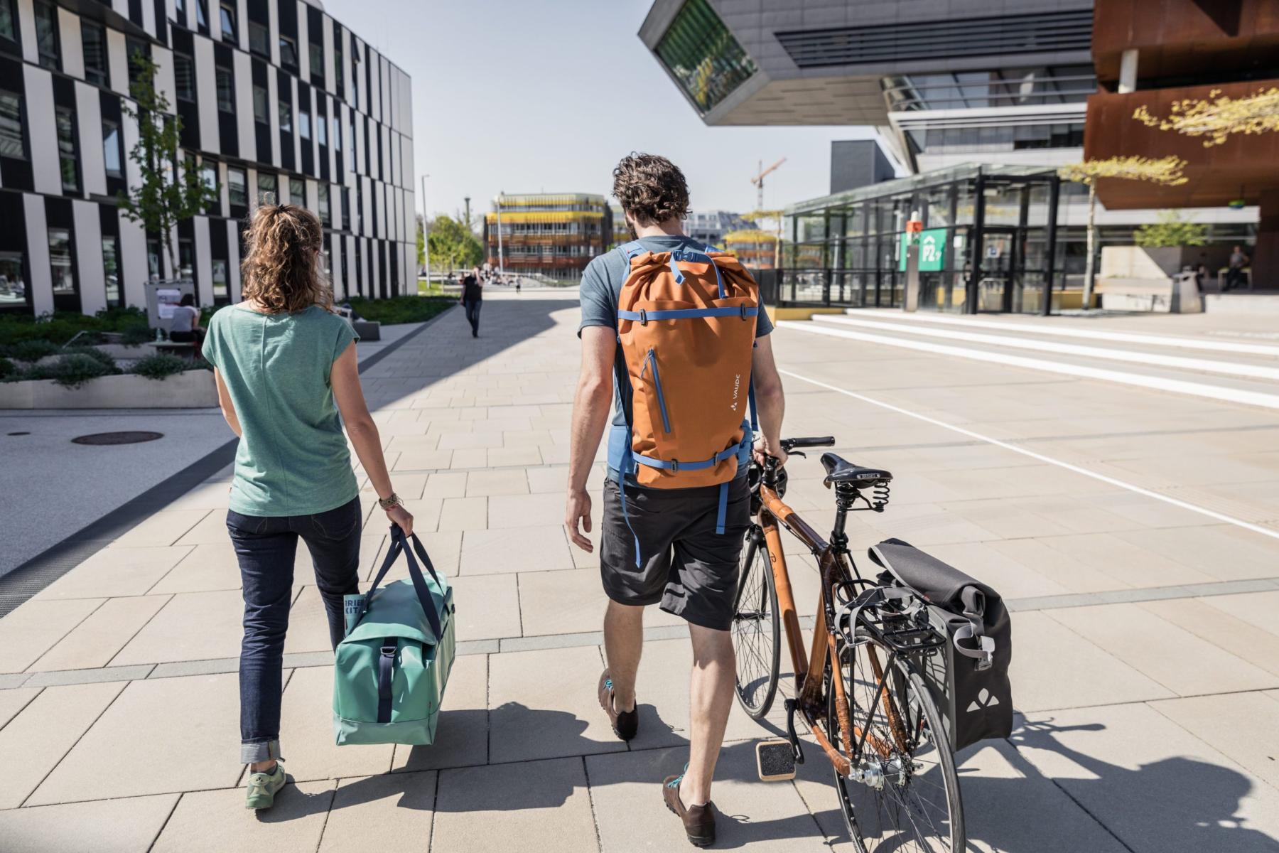Ein Mann mit einem VAUDE Rucksack auf dem Rücken schiebt ein City Fahrrad. Mit ihm läuft eine Frau mit einer Tasche in der Hand.