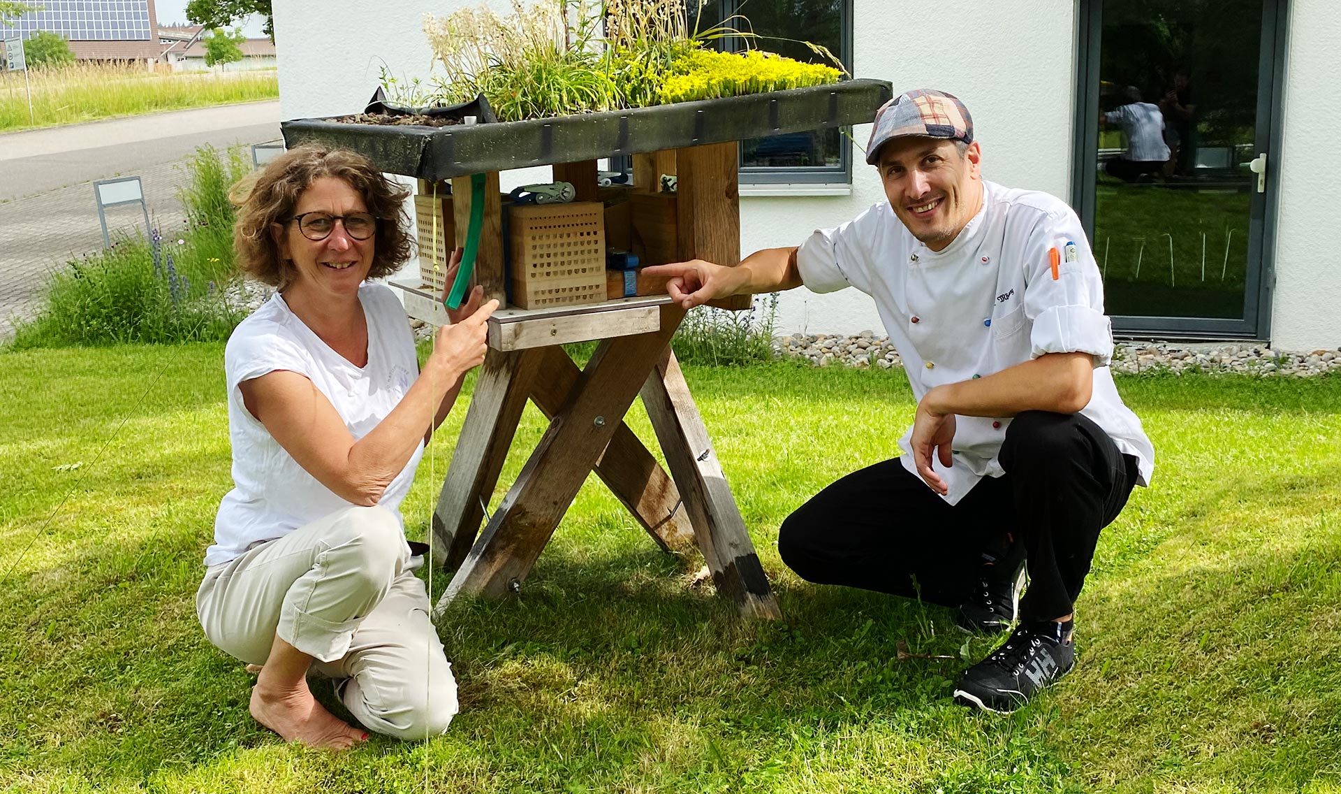 Eine Frau und ein Mann knien in Wiese neben einem Insektenhotel